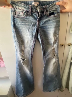 Blå bootcut jeans med slitningar - Coola ljusblå bootcut jeans med slitningar på framsidan och broderade bakfickor i rosa. Jeansens material är denim och de har klassisk femficksdesign med rosa detaljer längs sömmarna. Perfekt för dig som gillar en avslappnad och trendig look.