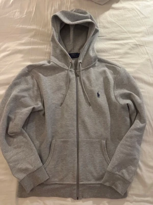Grå hoodie från Polo Ralph Lauren - Säljer en grå hoodie från Polo Ralph Lauren med dragkedja, huva med snören och två stora fickor framtill. Klassisk logga broderad på bröstet. Materialet är mjukt och bekvämt, perfekt för chill dagar. Stilen är enkel och tidlös.