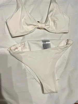 Vit bikini med knyt från H&M - Snygg vit bikini från H&M med knyt framtill på toppen. Både överdel och underdel har enkel, stilren design och är gjorda i mjukt syntetmaterial. Perfekt för sommarens strandhäng och poolpartyn.