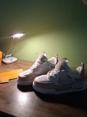 Louis Vuitton vita och beige sneakers - Snygga Louis Vuitton sneakers i vitt och beige med klassisk LV-logga på plösen och sidan. Skorna har en låg silhuett, robust sula och detaljer i mesh och läder. Perfekta för dig som vill ha en exklusiv och trendig look. Helt Nya men kommer tyvärr ej med originalbox.