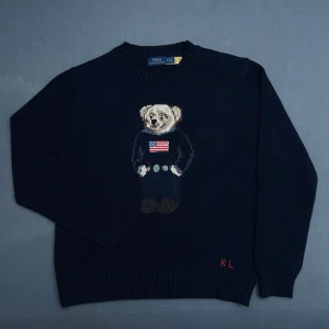 Mörkblå stickad tröja Polo Bear - Mörkblå stickad tröja från Polo Ralph Lauren med ikonisk Polo Bear på bröstet och  RL-broderi i rött vid nederkanten. Rund halsringning och ribbade muddar. Perfekt för dig som gillar klassisk preppy stil.