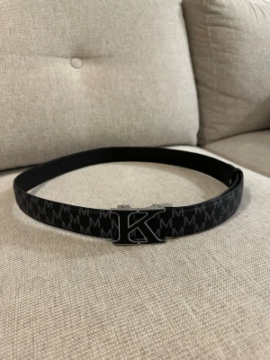 Svart bälte med K-logga  - Snyggt svart bälte med K-spänne och mönster av vita M-bokstäver längs hela bandet. Perfekt accessoar för att lyfta din outfit med en stilren och modern touch.