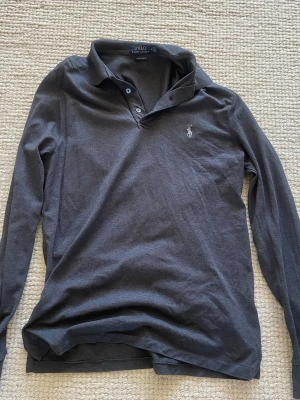 Mörkgrå långärmad piké från Polo Ralph Lauren - Snygg mörkgrå långärmad pikétröja från Polo Ralph Lauren i modellen Custom Slim Fit. Tröjan har klassisk krage, knäppning med två knappar och den ikoniska broderade loggan på bröstet. Tillverkad i mjukt bomullsmaterial för en bekväm känsla.