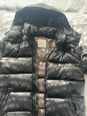 Svart dunjacka från Moncler - Snygg svart dunjacka från Moncler med glansig finish och stor huva. Jackan har en ficka med lock på ärmen och Moncler-logga. Dragkedja framtill och vadderad design som håller värmen under kalla vinterdagar. Perfekt för dig som vill ha både stil och funktion.