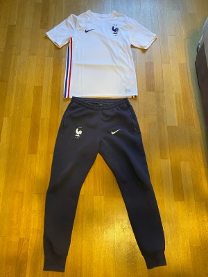 Frankrike Nike fotbollsträningsset - Säljer ett Frankrike fotbollsträningsset från Nike. Tröjan är vit med blå Nike-logga, landslagets emblem och ränder i blått, vitt och rött på sidan. Byxorna är mörkblå med resår i midjan och anklarna, samt Nike-logga och Frankrikes emblem. Perfekt för träning eller match.