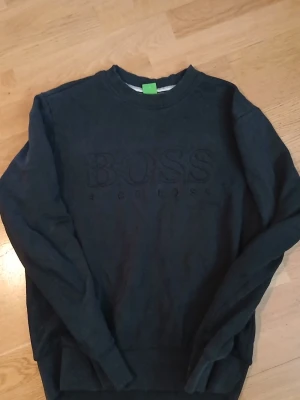 Svart sweatshirt från Hugo Boss - Svart sweatshirt från Hugo Boss med broderad logga på bröstet. Klassisk rund halsringning och ribbade muddar vid ärmslut och nederkant. Perfekt för en stilren och avslappnad look.
