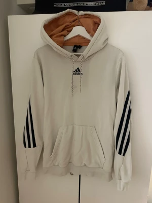Beige Adidas hoodie med svarta ränder - Snygg beige hoodie från Adidas med klassiska svarta ränder på ärmarna och svart logga på bröstet. Hoodien har en kontrastfärgad huva i brunt och mönstrade snören. Stor känguruficka framtill och ribbade muddar. Perfekt för en avslappnad streetwear-look. Finns en liten svag fläck på den borde enkelt gå bort med nå bra medel. Inget man lägger märke till dock.