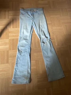 Ljusblå bootcut jeans med slitning - Ljusblå jeans med bootcut passform och klassisk femficksdesign. Jeansen har slitningar och hål på höger ben samt dragkedja och knapp framtill. Materialet är denim och färgen är ljusblå med viss tvättad effekt.