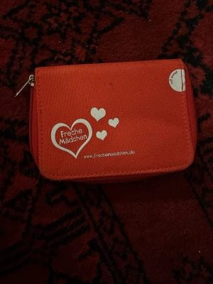 Röd plånbok Freche Mädchen - Snygg röd plånbok från Freche Mädchen i slitstark textil med vita hjärtdetaljer och logga på framsidan. Plånboken har dragkedja runtom och flera fack inuti, inklusive ett myntfack med egen dragkedja. Perfekt för att hålla ordning på kort och kontanter.