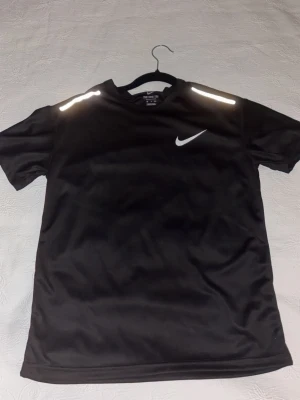 Svart Nike tränings T-shirt - Snygg svart tränings T-shirt från Nike med korta ärmar och vit logga på bröstet. Tröjan har reflekterande detaljer på axlarna och är tillverkad i ett lätt och snabbtorkande material som passar perfekt till träning och sport.