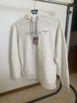 Vit hoodie från GANT med brodyr - Vit hoodie från GANT med broderad logotyp och amerikansk sköld på bröstet. Hoodien har huva med dragsko och ribbade muddar. Materialet är mjukt och bekvämt, perfekt för en avslappnad look.