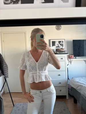 Vit blus med spets och volang - Säljer en vit blus med v-ringning, korta ärmar och broderad spets. Blusen har knappar framtill och en volang nedtill som ger en croppad look. Perfekt för dig som gillar romantiska detaljer och ljusa färger.