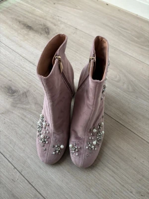Rosa stövletter med pärldekor - Säljer ett par rosa stövletter med pärldekorationer och små stenar på både fram- och sidorna. Skorna har en rund tå, blockklack och dragkedja på insidan. Materialet ser ut att vara mocka eller sammet, vilket ger en lyxig känsla.
