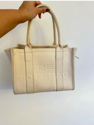Beige Tote Bag från Marc Jacobs - Säljer en beige Tote Bag från Marc Jacobs i kraftigt, strukturerat läder. Väskan har två rejäla handtag, präglad logga framtill och en stilren, boxig form. Perfekt för dig som gillar minimalistisk och trendig design.