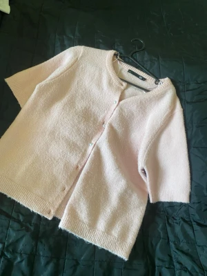 Ljusrosa kofta med pärlknappar - Ljusrosa kofta med korta ärmar och rund halsringning. Koftan har pärlknappar framtill och är stickad i ett mjukt material. Perfekt för att addera en söt touch till din outfit.