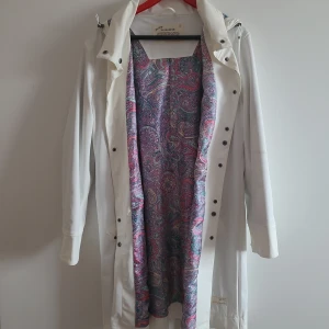 Regnkappa från Odd Molly - Säljer en vit regnkappa från Odd Molly med färgglatt paisleymönstrat foder i lila, rosa och blå toner. Kappan har huva, tryckknappar och långa ärmar. Perfekt för regniga dagar när du vill sticka ut lite extra.