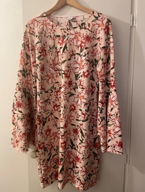 Snygg klänning med speciella ärmar, Gina Tricot - Säljer en blommig klänning med ljus bas och röda samt gröna detaljer. Klänningen har vida långärmar(volanger)  och en rak passform. Perfekt för dig som gillar ett romantiskt och färgstarkt uttryck. Materialet känns mjukt och lätt, troligen polyester. St: XL