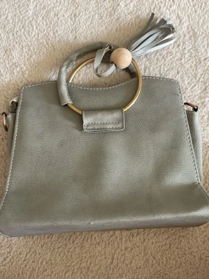 Grå handväska med guldring och tofs - Stilren grå handväska i slätt konstläder med en dekorativ guldfärgad ring som handtag och en snygg tofs med träpärla. Väskan har en enkel, boxig form och metallfästen på sidorna för axelrem. Perfekt för dig som gillar trendiga detaljer.