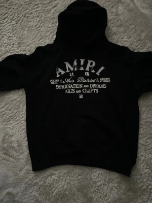Svart hoodie från Amiri med tryck - Svart hoodie från Amiri med vit texttryck på bröstet. Hoodien har klassisk huva och ribbade muddar. Materialet ser ut att vara mjukt och bekvämt, perfekt för en avslappnad och cool look. Trycket har en vintagekänsla med text om imagination och arts & crafts.