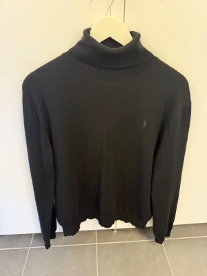 Svart polotröja från Polo Ralph Lauren stl L - Svart stickad polotröja från Polo Ralph Lauren med diskret broderad logga på bröstet. Tröjan har lång ärm och ribbade muddar vid hals, ärmslut och nederkant. Perfekt för lager-på-lager och en clean look.