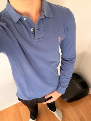 Polo Ralph Lauren Piké Långärmad - Klassisk mörkblå långärmad pikétröja från Polo Ralph Lauren med två knappar och rosa broderad logga på bröstet. Tillverkad i mjuk bomull och har en stilren krage. Perfekt för dig som gillar en enkel men snygg look. Finns inga defekter.