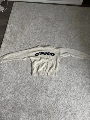 Beige stickad tröja  - Stickad beige tröja med svarta lucky motiv över bröstet. Tröjan har rund hals, långa ärmar och ribbade muddar vid ärmslut och nederkant. Perfekt för dig som gillar en lekfull och trendig stil.