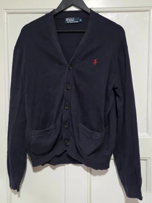 Mörkblå kofta från Polo Ralph Lauren - Mörkblå kofta från Polo Ralph Lauren med klassisk röd logobrodyr på bröstet. Koftan har v-ringning, knäppning med fem knappar och två fickor framtill. Materialet är mjukt och tunt, perfekt för lager-på-lager. Snygg och enkel att matcha med olika outfits.