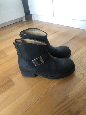 Svarta boots med spänne Johnny bulls  - Snygga svarta boots i läder med grov klack och coolt metallspänne på sidan. Bootsens insida är ljust fodrad och de har en robust sula som passar perfekt till jeans eller kjol. Klassisk och tidlös design som funkar till många olika stilar.
