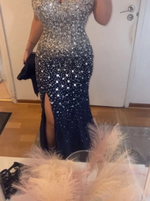 Glittrig långklänning med slits - Säljer en elegant långklänning med glittriga paljetter upptill och marinblå nederdel. Klänningen har v-ringning och en snygg slits framtill som ger en cool touch. Perfekt för dig som vill sticka ut och glänsa på balen eller festen!