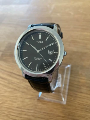 Timex Indiglo Date WR30 38mm Herrklocka - Elegant Timex Indiglo herrklocka med en klassisk design. Klockan har en svart urtavla med tydliga markeringar och datumvisning. Boetten är i silverfärg och kompletteras av ett svart läderarmband. Indiglo-funktionen ger en praktisk bakgrundsbelysning. Vattentålig till 30 meter (WR30M). Modellnummer T21752. Mycket fint skick enligt bilder, inga repor. Behöver bara nytt batteri.  Nypris: 900 SEK Begagnat värde: < 600 SEK Mitt pris: 489 SEK  Skickas alltid snabbt och spårbart. Skriv vid frågor och s
