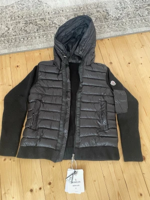 Moncler cardigan - Snygg svart cardigan från Moncler med stickade svarta ärmar och huva. Den har två framfickor med knappar, dragkedja framtill och en Moncler-logga på ärmen. Materialet är en kombination av dun och stickad textil för en modern look. Den är i väldigt bra skick och priset kan diskuteras 