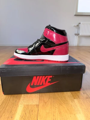 Nike Air Jordan 1 svart/röd high - Säljer ett par Nike Air Jordan 1 high sneakers i svart och rött med blankt, patenterat läder och vit sula. Klassisk siluett med ikonisk Swoosh och högt skaft. Perfekt för dig som vill sticka ut med en tidlös och trendig modell.