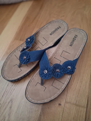 Blå blommiga sandaler Fashion - Snygga blå sandaler med blommiga detaljer och små nitar framtill. Modellen är en klassisk flip-flop med beige sula och dekorativa sömmar runt kanten. Perfekta för sommaren och lätta att matcha med olika outfits.