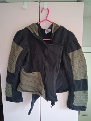 hoodiejacka med huva från Zapata - Unik hoodiejacka i patchwork-stil med stora paneler i olivgrönt och svart. Jackan har en stor, asymmetrisk huva och dragkedja framtill. Tillverkad i mjukt bomullstyg med vadderade detaljer på ärmarna och midjebälte. Perfekt för dig som gillar alternativ stil.