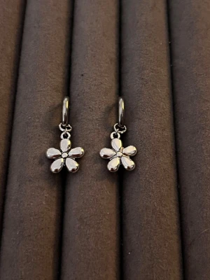 Blommiga örhängen i silverfärg - Säljer ett par söta örhängen i silverfärg med hängande blommor. Blommorna har fem rundade kronblad och en liten detalj i mitten. Örhängena är i metall och har en enkel, stilren design som passar till många olika looks.