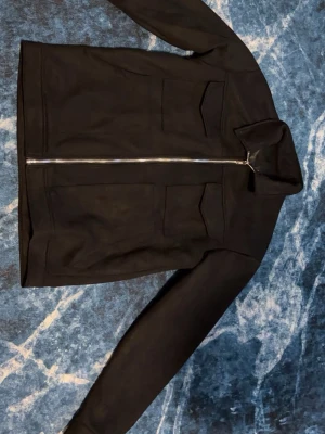 Mockajacka  - Stilren svart overshirt med rak passform och två stora bröstfickor. Jackan har en klassisk krage och hel dragkedja framtill. Materialet ser ut att vara mjukt och lite tjockare, perfekt för lager-på-lager. Passar dig som gillar minimalistisk och clean stil.