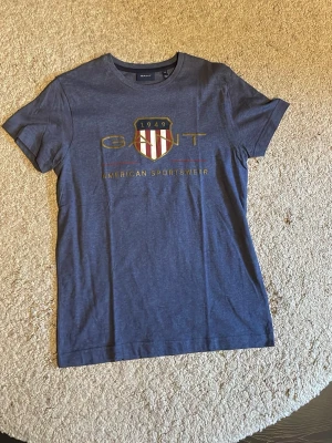 Blå GANT t-shirt med tryck XS - Snygg blå t-shirt från GANT med klassiskt tryck framtill och sköld med rödvita ränder. Modellen har rund hals och korta ärmar. Tillverkad i mjuk bomull som känns skön mot huden. Perfekt för en avslappnad och sportig stil.