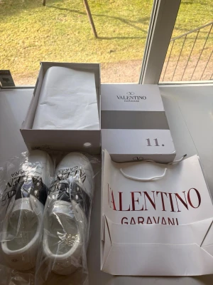 Valentino Garavani vit sneakers med svart rand  - Snygga vita sneakers från Valentino Garavani med svart band över mitten och ikoniska nitar på sulan. Skorna är låga och har en stilren design i läder med vita snören. Perfekta för dig som vill ha en exklusiv touch till din outfit.