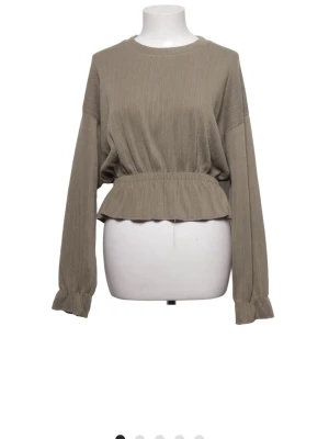 Beige ribbad topp med volang - Trendig beige topp med ribbad struktur och volang i midjan. Långärmad modell med rund hals och resår i midjan för en snygg passform. Köpt från Sellpy 
