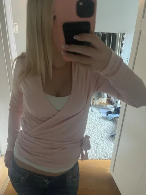 Ljusrosa omlottopp med knyt - Söt ljusrosa omlottopp med knyt i sidan och lång ärm. Toppen har en djup v-ringning och är i mjukt, stretchigt material. Perfekt att styla med jeans eller kjol för en trendig look.