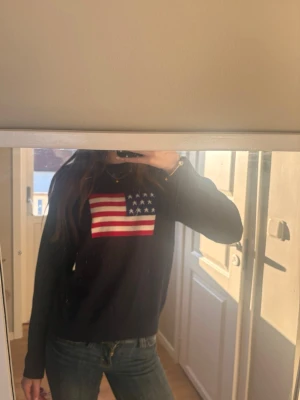 Mörkblå tröja med USA-flagga RYVLS - Snygg mörkblå tröja från RYVLS Stockholm med amerikansk flagga tryckt på bröstet. Tröjan har rund halsringning och ribbade muddar vid ärmslut och nederkant. Perfekt för dig som gillar stilrena plagg med en twist.