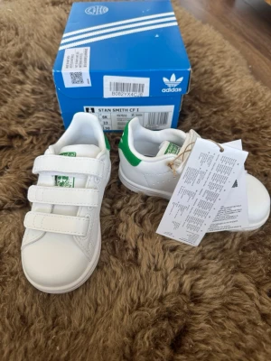 Adidas Stan Smith vita sneakers - Klassiska vita Adidas Stan Smith sneakers med gröna detaljer på hälen och tungan. Skorna har tre kardborreband för enkel påtagning och är tillverkade i syntetmaterial med gummisula. Perfekta för en clean och sportig look.