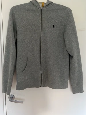 Grå kofta från Polo Ralph Lauren - Snygg grå zip-hoodie från Polo Ralph Lauren med klassisk logga broderad på bröstet. Hoodien har dragkedja framtill, två fickor och en mjuk bomullsblandning. Perfekt för en chill och avslappnad stil. Det är den största barnstorleken vilket motsvarar ungefär S men passar XS och M beroende på hur man vill ha passformen. Möts i Stockholm vid försäljning.