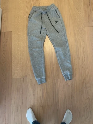 Nike tech byxor  - Snygga gråa mjukisbyxor från Nike med svarta detaljer och dragsko i midjan. Byxorna har smal passform, muddar vid bensluten och två fickor med dragkedja. Perfekta för chill eller träning.