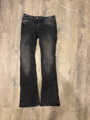 Svarta bootcut jeans med rosetter - Säljer ett par svarta bootcut jeans med snyggt slitna detaljer och dekorativa rosetter på bakfickorna. Bakfickorna är inte sånna med knäppe utan man kan lägga ner ex en telefon som med vanliga fickor 