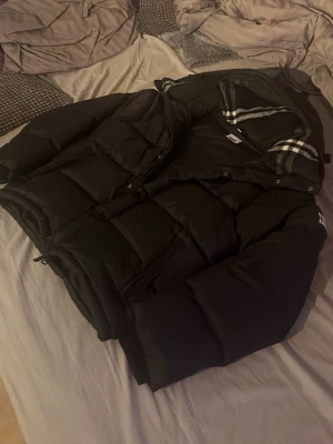 Svart burberry jacka med avtagbara armar  - Säljer en svart burberry jacka med avtagbara armar. Jackan har två stora fickor med dragkedja och knappar framtill. Perfekt för lager-på-lager och håller dig varm under kyliga dagar. Snygg och stilren design som passar till många outfits.