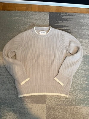 Maison Margiela Hoodie  - Snygg beige stickad tröja från Maison Margiela med rund halsringning och ribbade muddar. Tröjan har en stilren design med diskreta vita detaljer vid ärmslut och nederkant. Perfekt för lager-på-lager och enkel att matcha med olika outfits.