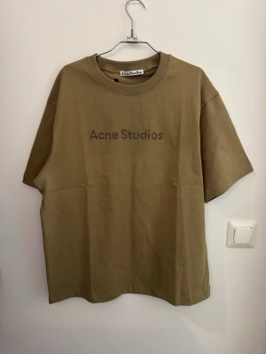 Beige Acne Studios t-shirt - Snygg beige t-shirt från Acne Studios med logga i små pärlor på bröstet. Klassisk rund halsringning och avslappnad passform. Perfekt till jeans eller shorts för en clean och trendig look.