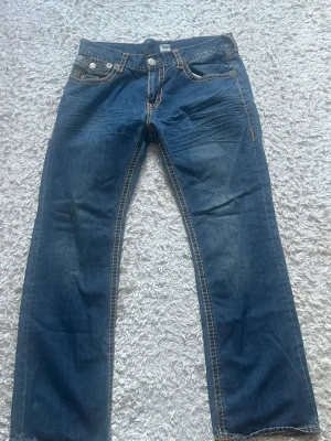 Blå True Religion jeans med kontrastsömmar - Säljer ett par blå jeans från True Religion med raka ben och tydliga kontrastsömmar. Jeansen har klassiska fem fickor, vita knappar och snygga detaljer på bakfickorna med True Religion-logga. Materialet är denim i bomull.
