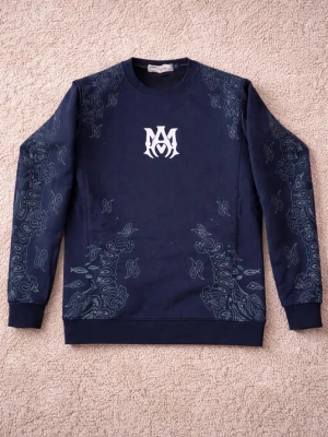 SVART sweatshirt från AMIRI - Svart sweatshirt från AMIRI med vit AMIRI-logga centrerad framtill och gråa mönstrade paneler längs ärmarna och nederkanten.  Plagget är mycket sparsamt använt och i nyskick. Inga fläckar, noppor eller sprickor i trycket. Färgen är jämn och materialet har behållit sin form. Normal passform och bekväm kvalitet som passar till många tillfällen.  Säljs då den inte kommer till användning.